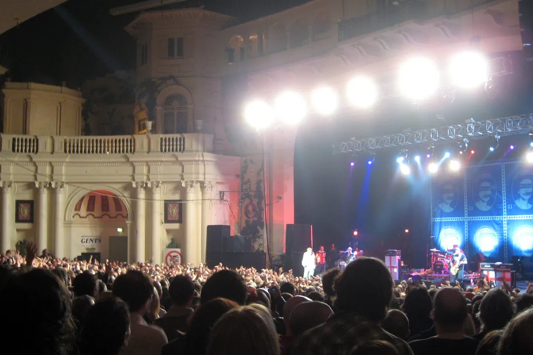 The Sex Pistols on stage at Brixton Academy in 2007, lit white and blue, under the venue's ornate proscenium, a packed crowd in front.