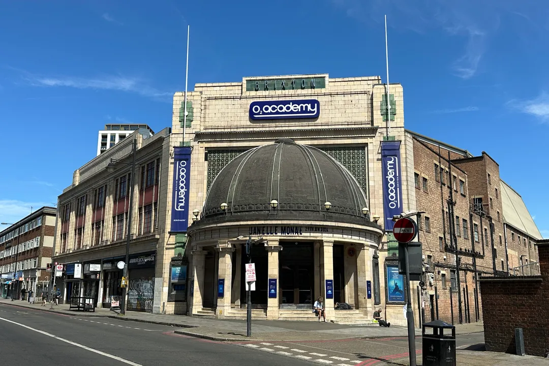 Brixton Academy's domed Art Deco front on a sunny day in June 2024, with O2 Academy banners and a quiet Stockwell Road in front.